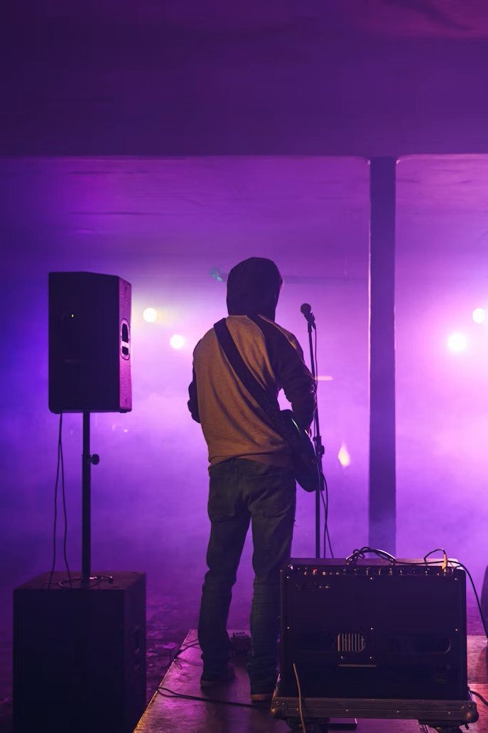 Musicien de dos sur une scène, entre une enceinte sur pied et un ampli, sous des lumières violettes